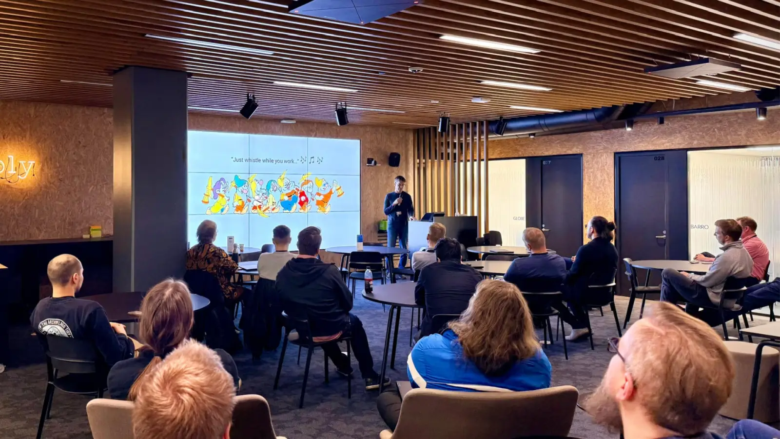 Teemu Piirainen presenting to a seated audience at Elisa, beside a slide reading 'Just whistle while you work' with the seven dwarfs.